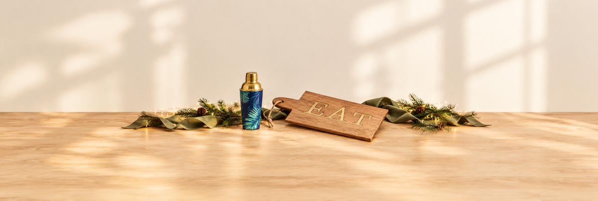 Palm leaf cocktail shaker and mango wood EAT serving board with olive ribbon and festive greenery.