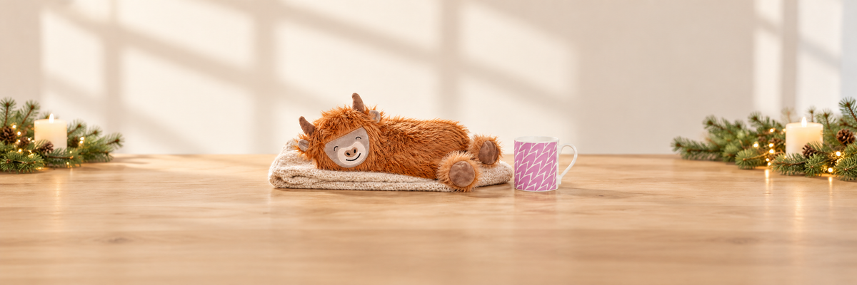 Highland Cow hot water bottle on a knitted throw beside a pink leaf mug and festive greenery.