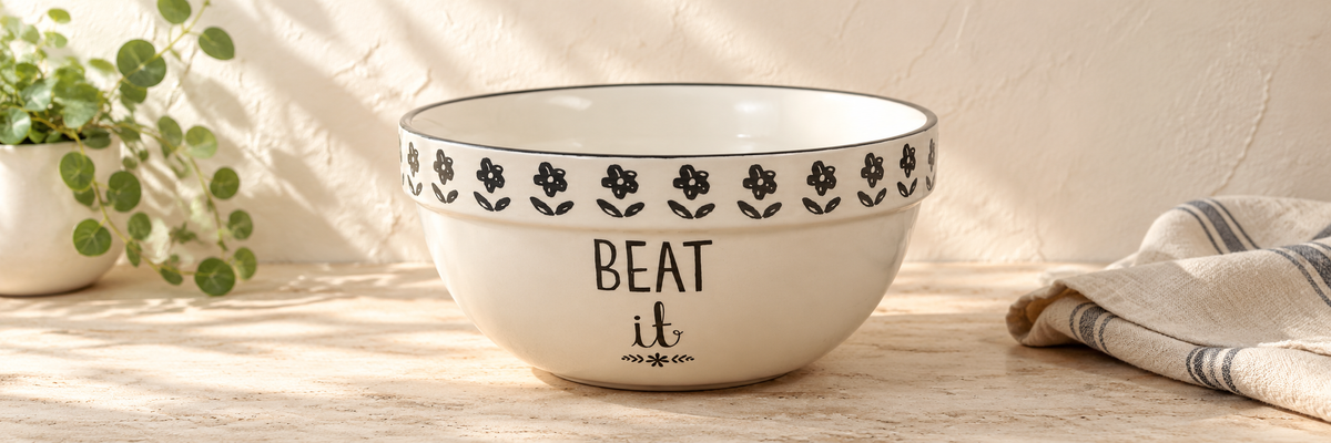 Cream "Beat it" mixing bowl with a black floral border on a softly lit countertop.