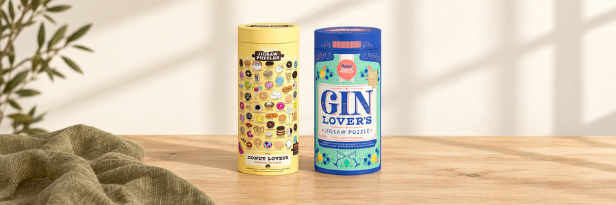 Donut Lover’s and Gin Lover’s jigsaw puzzle tubes on a pale wooden table